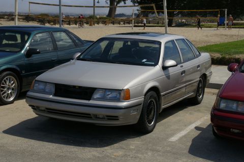 1990 Ford Taurus - Silver 1990 Ford Taurus parked outdoors with other vehicles and a beach volleyball court in the background.
