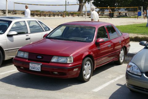 1990 Ford Taurus - A red 1990 Ford Taurus is parked in a lot near a beach, with people playing volleyball in the distance.