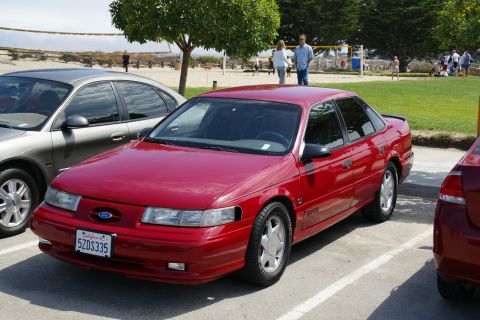 1990 Ford Taurus - A red sedan is parked next to another car with a beach and volleyball court in the background.