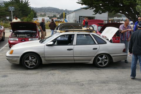 1990 Ford Taurus - A group of people gather around cars parked outdoors, with one sedan prominently displayed with a roof rack.