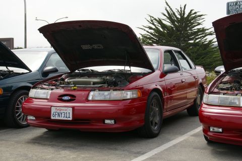 1990 Ford Taurus - A red 1990 Ford Taurus SHO is parked with its hood open, revealing the engine bay.