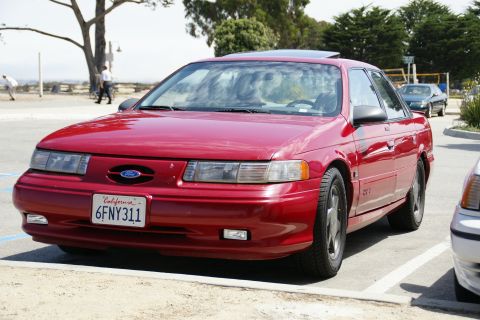 1990 Ford Taurus - Red 1990 Ford Taurus sedan parked in a lot with another vehicle partially visible.