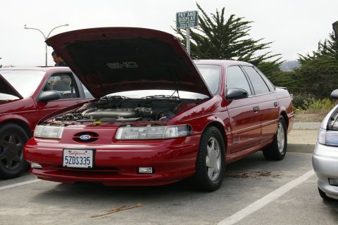 1990 Ford Taurus - A red Ford Taurus SHO is parked with its hood open, revealing the engine compartment. Another Taurus and a silver car are visible.