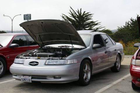 1990 Ford Taurus - A silver 1990 Ford Taurus sedan is parked with its hood open, revealing the engine compartment. Another red car is visible to the left.