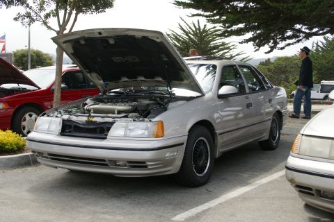 1990 Ford Taurus - A silver sedan, identified as a 1990 Ford Taurus SHO, is parked with its hood open, revealing the engine compartment.