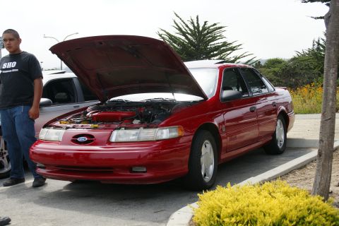 1990 Ford Taurus - A red 1990 Ford Taurus sedan is parked with its hood open, showing the engine compartment.