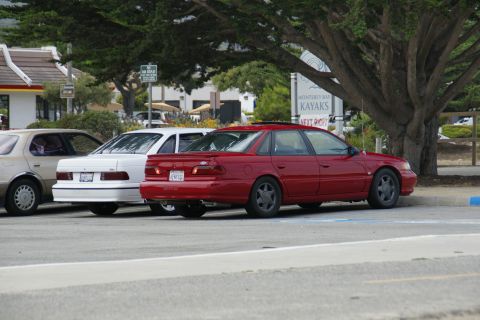 1990 Ford Taurus - A red sedan is parked in a lot next to other vehicles, with a sign for Monterey Bay Kayaks visible.