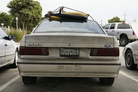 1990 Ford Taurus - A beige 1990 Ford Taurus SHO is parked, with a surfboard strapped to its roof rack.