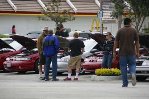1990 Ford Taurus - Several people gather around cars with hoods up at a car meet.