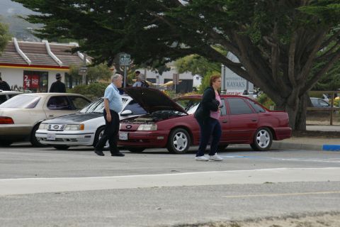 1990 Ford Taurus - A red 1990 Ford Taurus sedan with its hood open is parked next to another car, with people walking by.