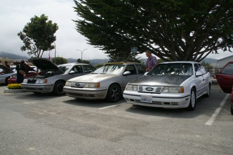 1990 Ford Taurus - Multiple Ford Tauruses are parked in a lot, with two of them having their hoods open.