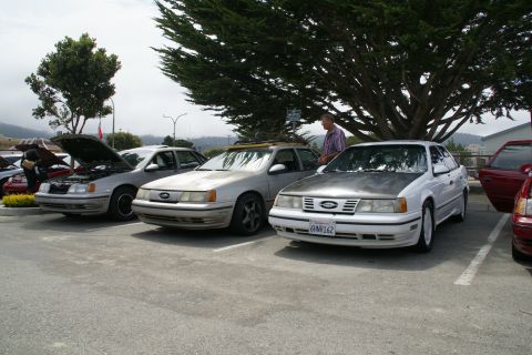 1990 Ford Taurus - A white 1990 Ford Taurus with a matte hood is parked next to other vehicles, with a man standing nearby under a large tree.