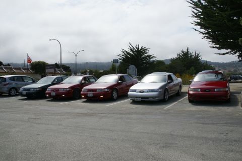 1990 Ford Taurus - A row of Ford Taurus sedans, mostly red, parked in a lot under a cloudy sky.