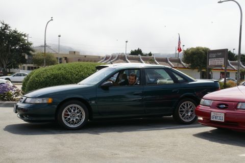 1990 Ford Taurus - A man smiles from inside a green sedan, parked next to a red car with a California license plate.