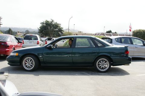 1990 Ford Taurus - A dark green sedan is parked in a lot, with other vehicles visible in the background.