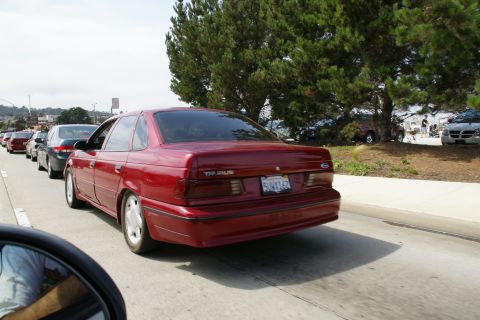 1990 Ford Taurus - A red 1990 Ford Taurus is seen from behind in a line of cars on a sunny day.