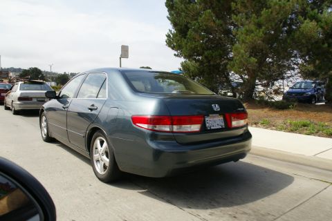 1990 Ford Taurus - A dark gray sedan is seen from the rear in traffic, with other vehicles and trees in the background.