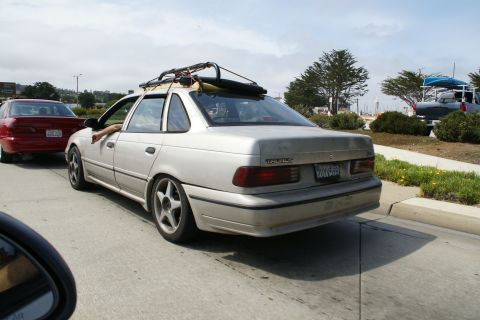1990 Ford Taurus - A silver 1990 Ford Taurus sedan with a roof rack is seen from the rear, driving on a sunny day.