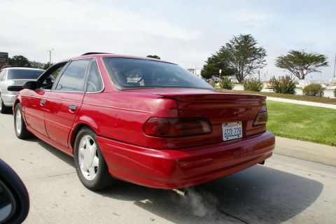 1990 Ford Taurus - Rear view of a red 1990 Ford Taurus sedan driving on a road, with another car behind it.