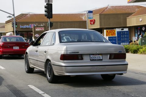 1990 Ford Taurus - A silver 1990 Ford Taurus SHO drives down the road, with a red Ford sedan visible in the background.