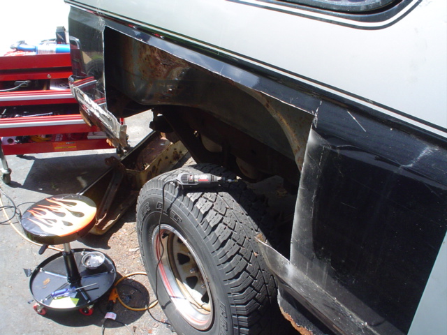 close-up of rusted rear quarter panel of a vehicle being worked on, with a tire and tool cart visible