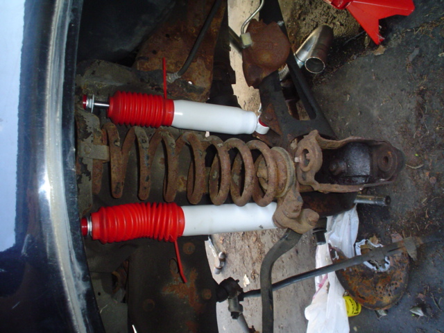 Rusty coil springs and white shocks with red bellows are mounted to a vehicle's suspension assembly