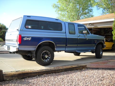 1996 Ford F-250 - This blue Ford pickup truck, equipped with a camper shell, is parked in a driveway. A yellow car is visible in the garage behind it.