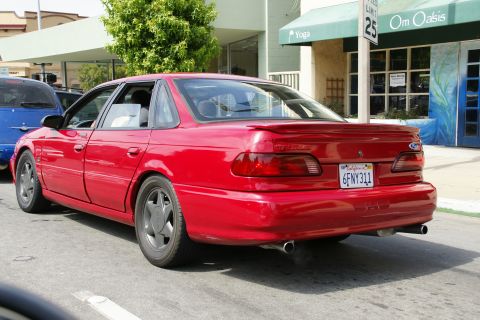 1990 Ford Taurus - A red sedan is parked on a street, with a blue vehicle visible to its left.