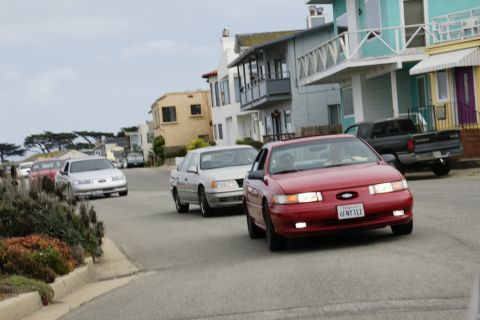 1990 Ford Taurus - Red 1990 Ford Taurus driving down a street with other vehicles and houses visible.