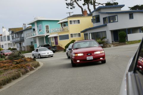 1990 Ford Taurus - A red Ford Taurus drives down a residential street past colorful houses.
