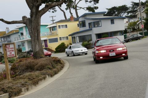 1990 Ford Taurus - A red 1990 Ford Taurus drives down a residential street, with other vehicles and houses in the background.