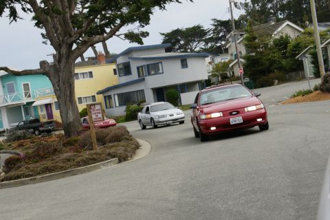 1990 Ford Taurus - A red 1990 Ford Taurus drives down a residential street, with another car visible in the background.