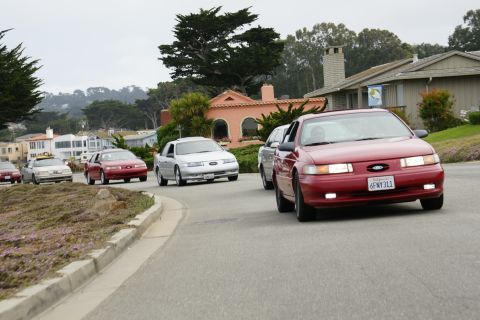 1990 Ford Taurus - A line of cars, including a red Ford Taurus, drives down a suburban street.