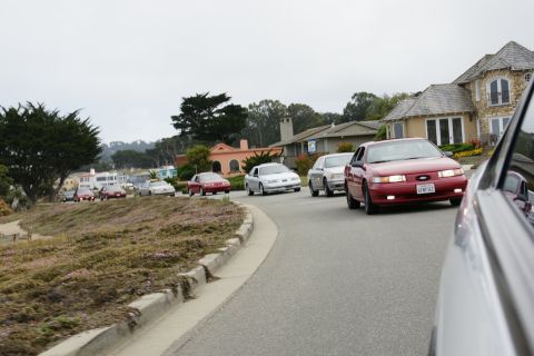1990 Ford Taurus - A line of cars, including a red 1990 Ford Taurus, drive down a suburban street with houses and trees.
