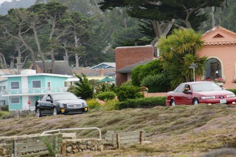 1990 Ford Taurus - Two cars drive along a scenic road lined with houses and lush vegetation.