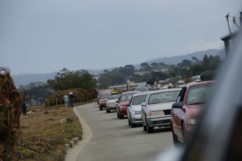 1990 Ford Taurus - A line of cars, including a 1990 Ford Taurus, travels along a coastal road with houses and hills in the distance.