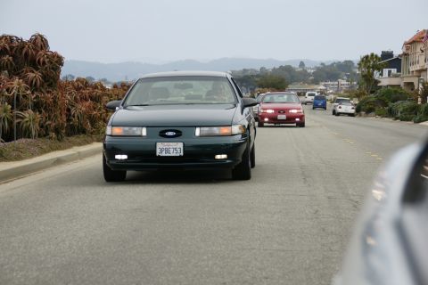 1990 Ford Taurus - A dark green 1990 Ford Taurus drives down a coastal road, with other vehicles visible ahead.