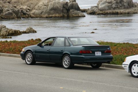1990 Ford Taurus - Rear view of a 1990 Ford Taurus sedan parked on a coastal road with a rocky shoreline in the background.