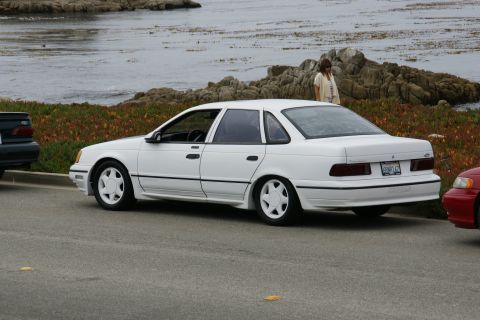 1990 Ford Taurus - A white sedan is parked on the side of the road with a rocky coastline and ocean in the background.