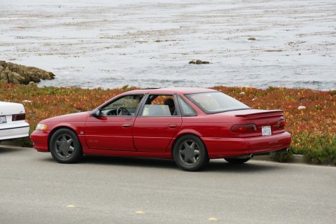 1990 Ford Taurus - A red sedan is parked on the side of the road with the ocean in the background.