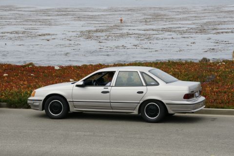 1990 Ford Taurus - A silver 1990 Ford Taurus sedan is seen from the side, parked near a coastal area with the ocean in the background.