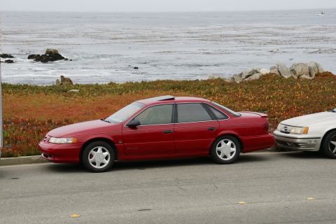 1990 Ford Taurus - A red sedan is parked by the ocean, with another vehicle beside it.