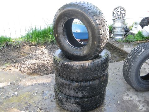 stack of four off-road tires, one spare tire to the right, and a stack of rims in the background