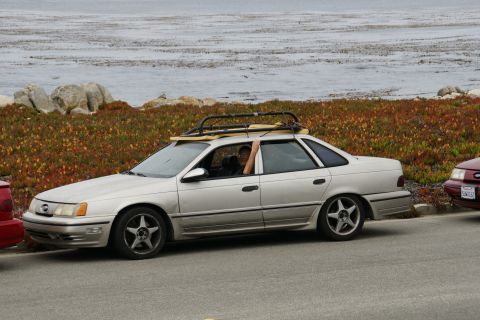 1990 Ford Taurus - A silver 1990 Ford Taurus sedan is parked near the ocean, equipped with a roof rack and a surfboard.