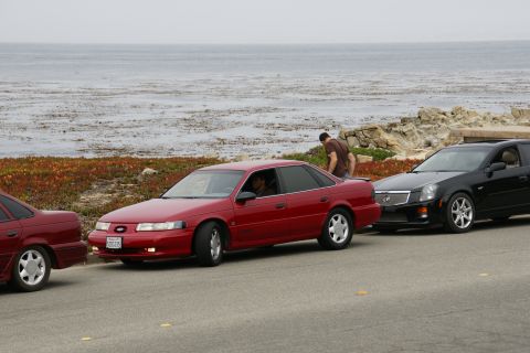 1990 Ford Taurus - A red sedan is parked on the side of the road with the ocean in the background.