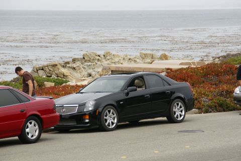 1990 Ford Taurus - A black Cadillac sedan is parked near the coast, with a red car to its left and the ocean in the background.