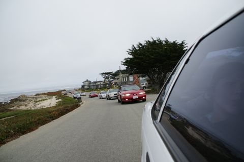1990 Ford Taurus - A line of cars, including a red 1990 Ford Taurus, drive along a scenic coastal highway.