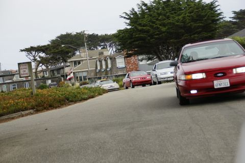 1990 Ford Taurus - Red 1990 Ford Taurus leads a line of cars on a street with coastal buildings and trees.