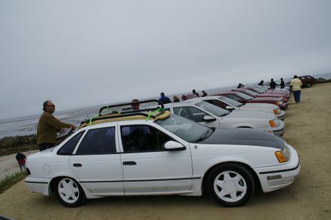 1990 Ford Taurus - A line of white and red sedans are parked on a sandy area near the ocean, with people milling around.