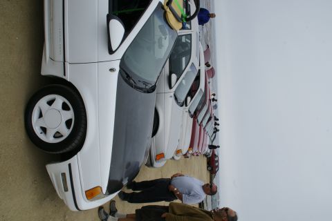 1990 Ford Taurus - A white 1990 Ford Taurus with a gray hood is parked in a line of vehicles, with two men standing nearby.